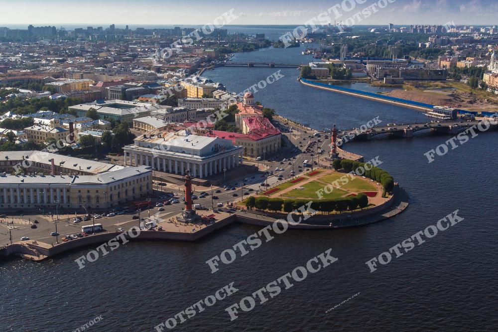 На изображении представлена Стрелка Васильевского острова, одно из символичных мест Санкт-Петербурга. В центре внимания — здание Биржи и две Ростральные колонны, которые зажигаются в праздничные дни. На заднем плане виден мост, соединяющий Васильевский остров с другими частями города, и широкая гладь реки Нева. Панорамный вид сверху подчеркивает величественную архитектуру и масштаб этого исторического района, насыщенного культурными и архитектурными памятниками.